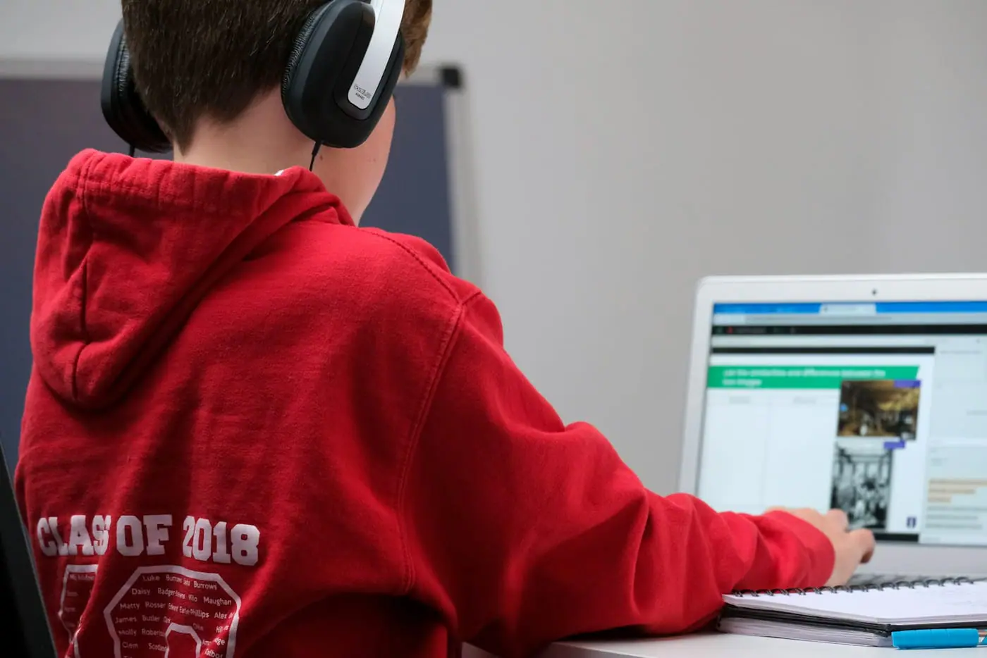 A boy in a red hoodie and headphones follows a lesson on a laptop