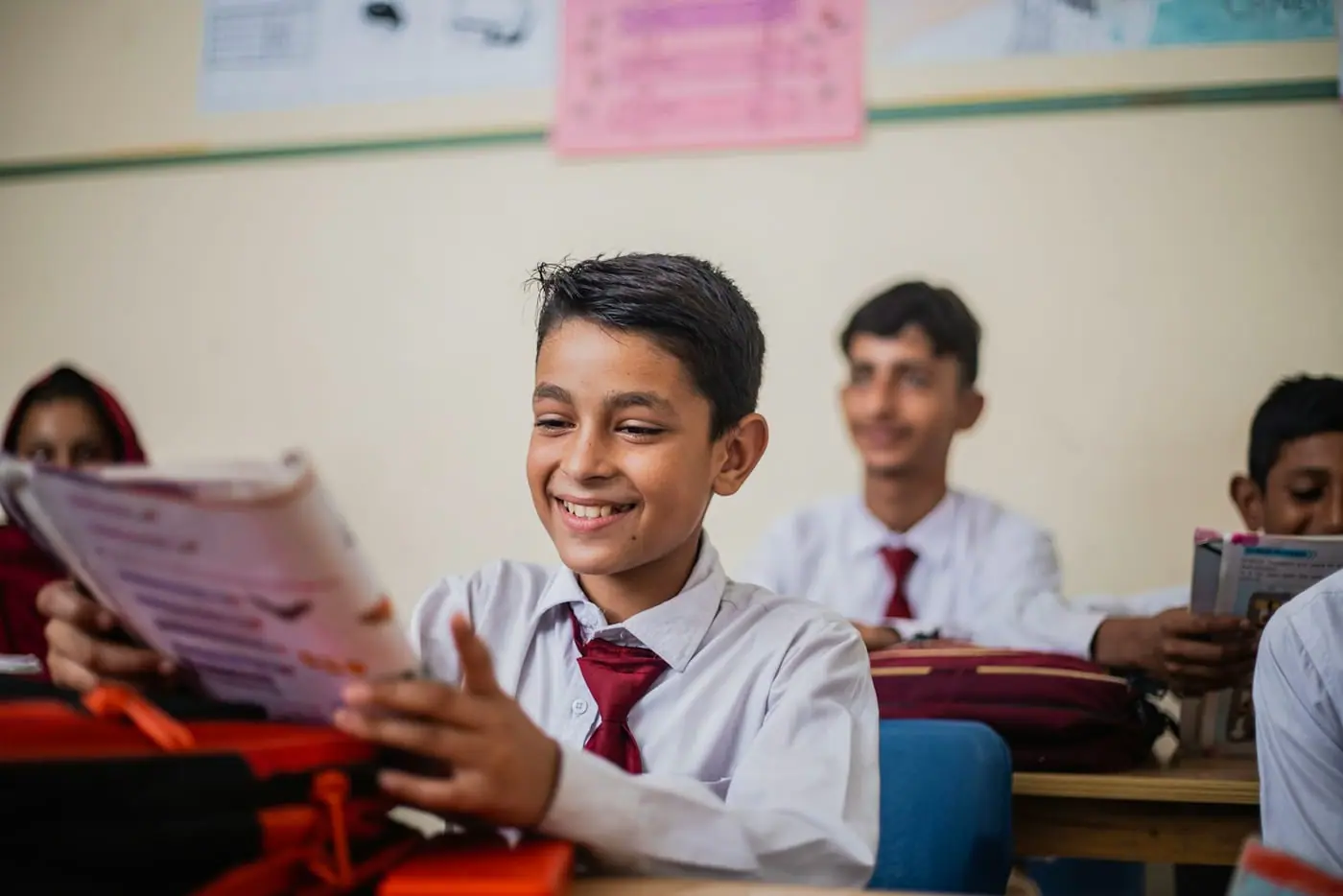 Schoolboys in white uniforms and red ties smile over an open book in a classroom in Pakistan