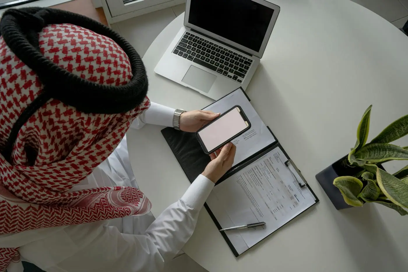 Seen from above, a student in a white thobe and red ghutra works at a desk with a laptop, notebook and phone