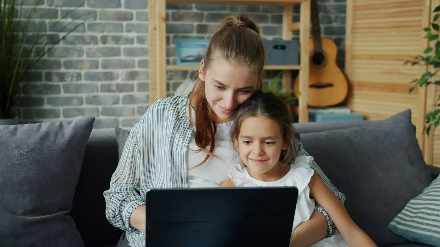 A mother and her daughter look at a laptop together on a sofa