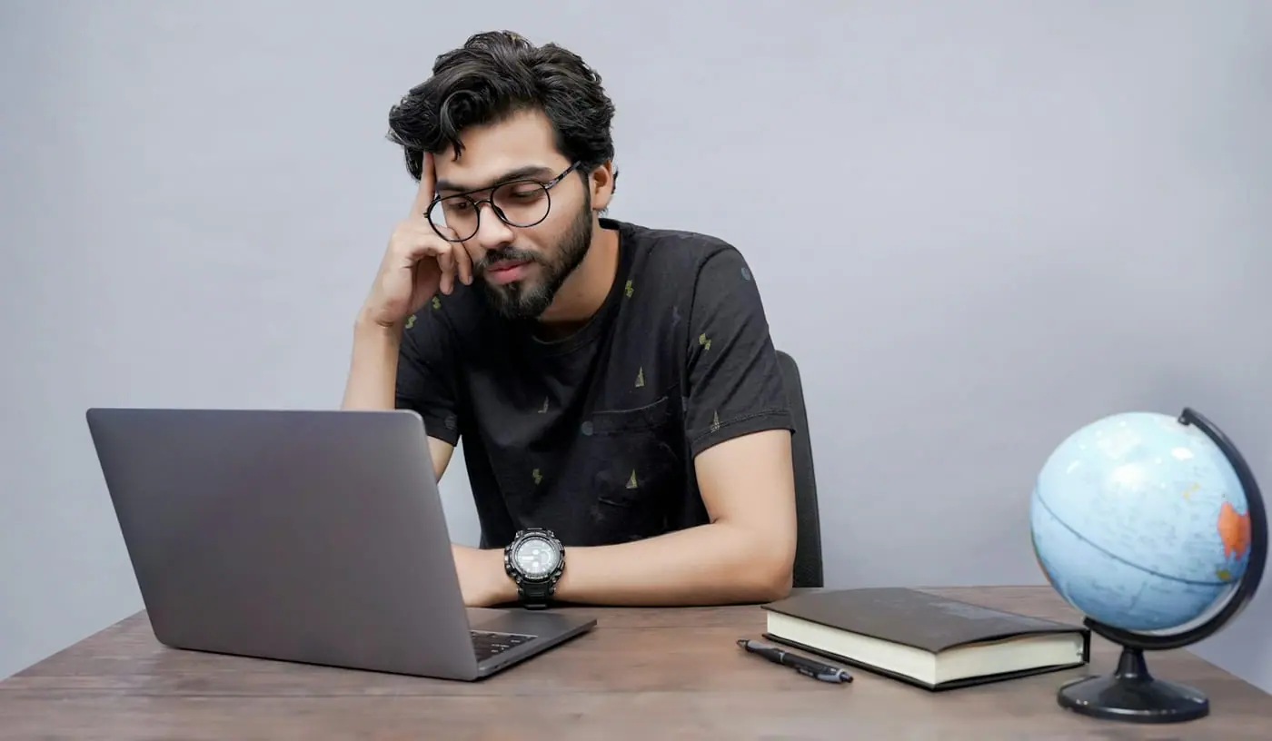 A young man studies at a laptop at a wooden desk with a globe and textbooks