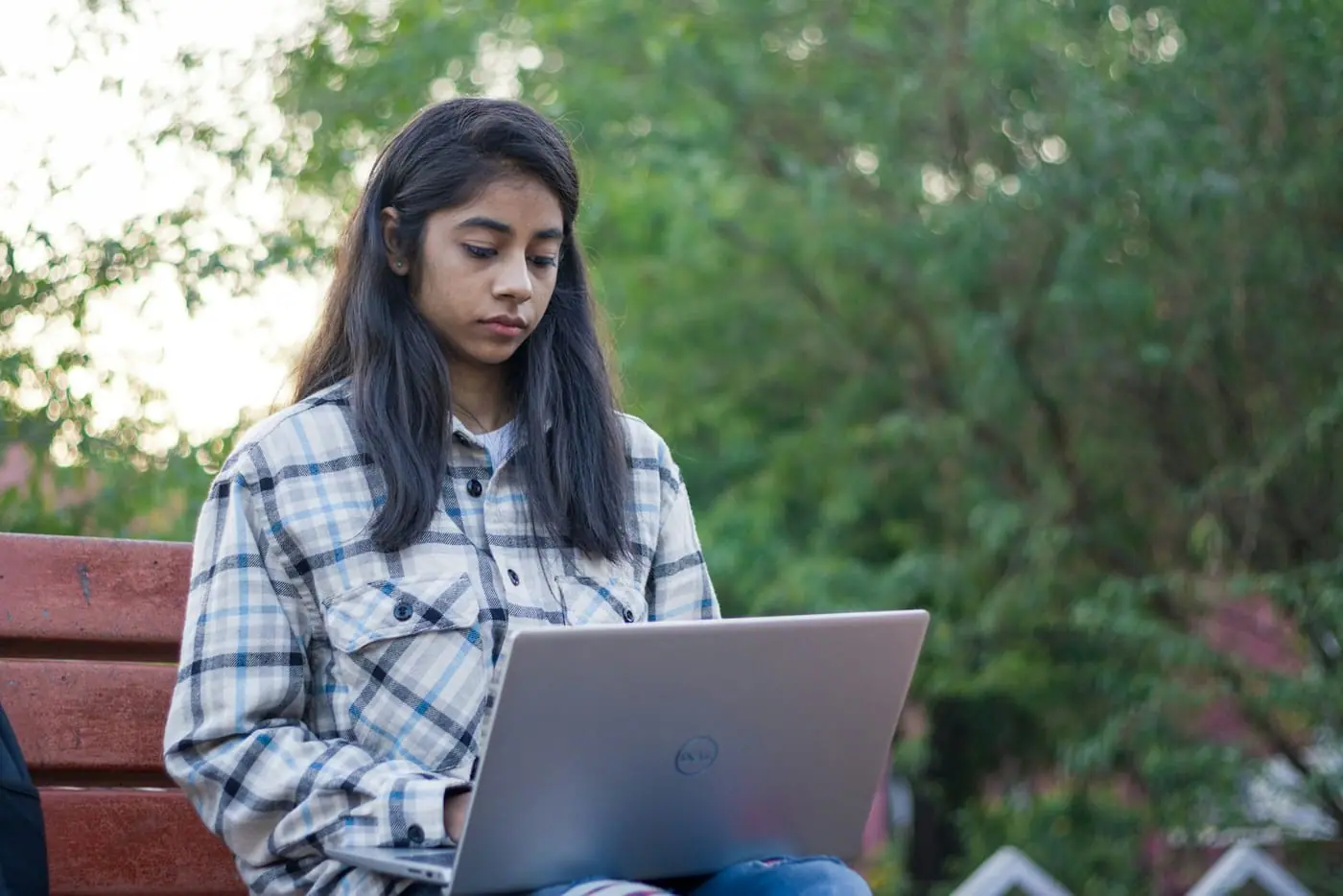 A teenage girl sits on a park bench with a laptop open on her knees