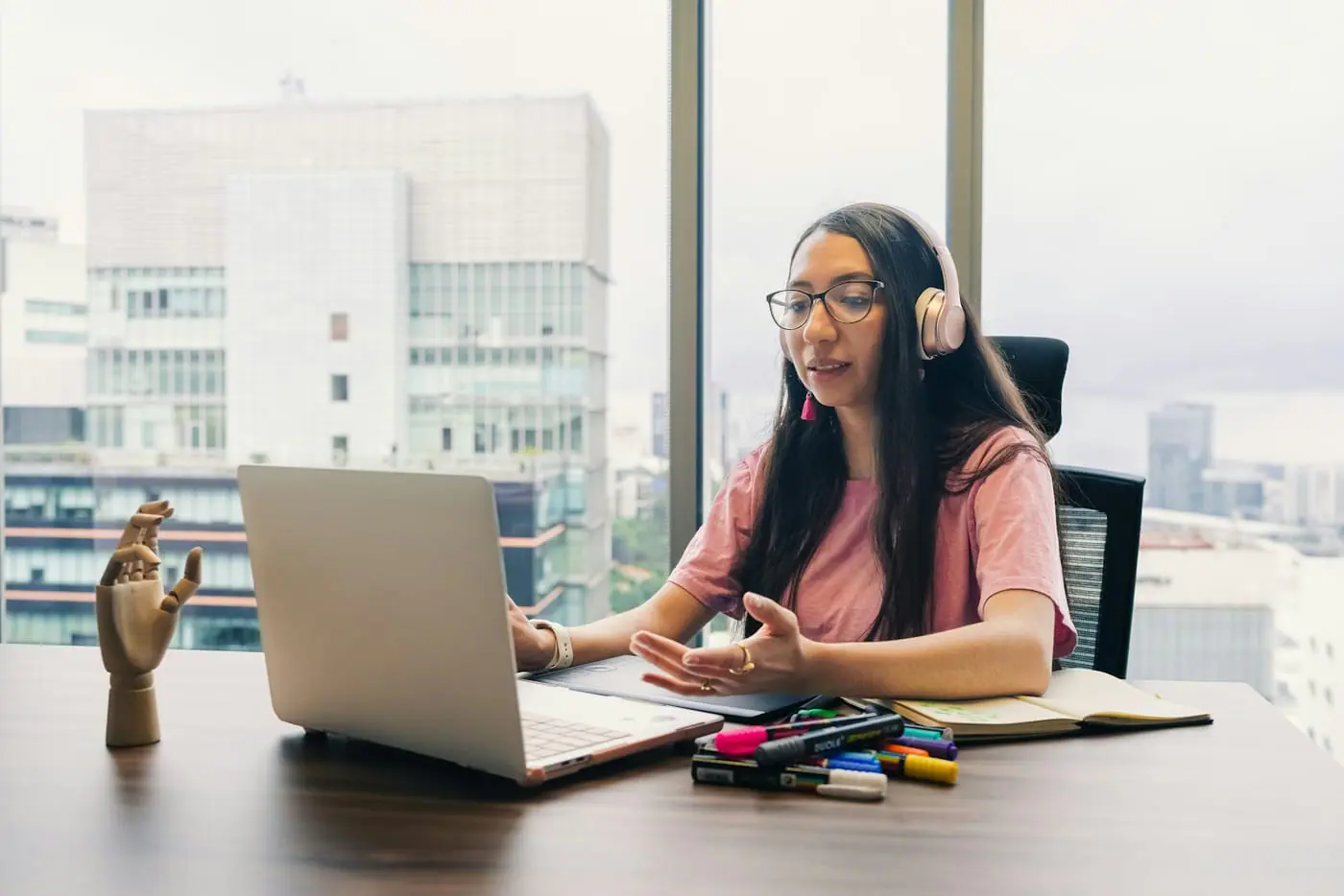 A student with headphones talks to a tutor on a laptop video call beside a window
