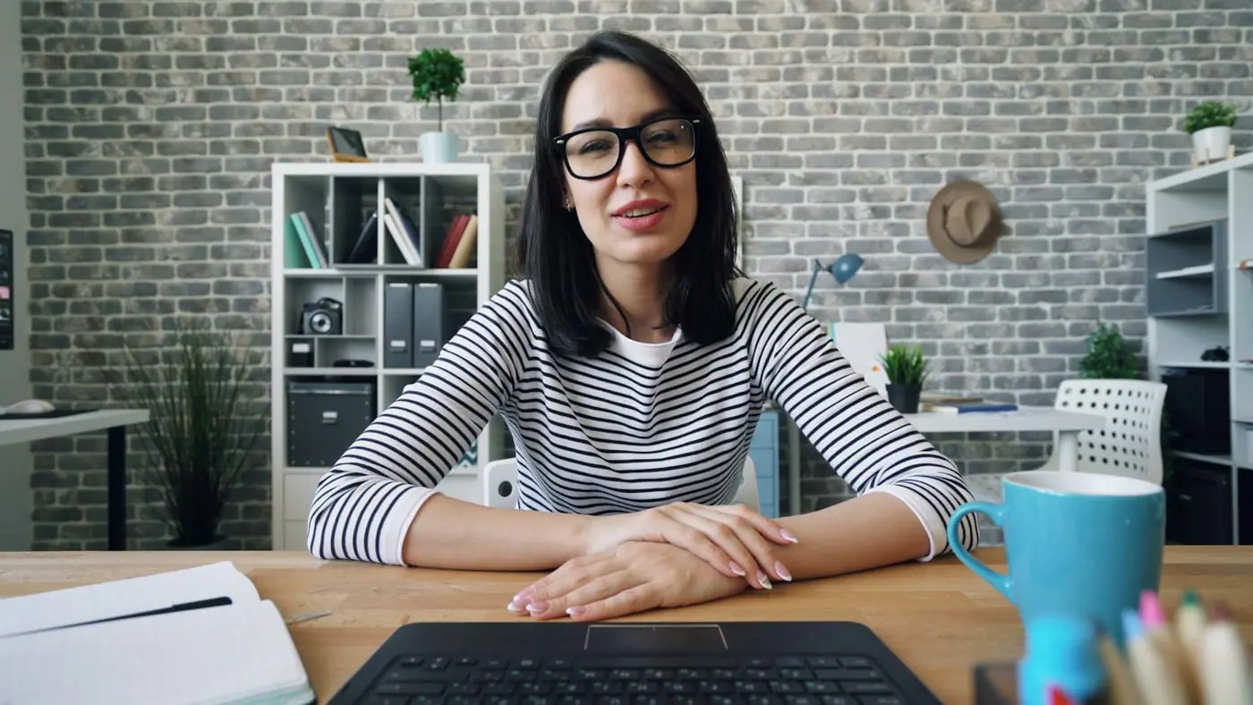 A teacher with glasses sits at a desk facing the camera, as seen on a video call